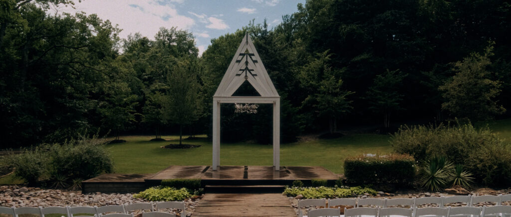 Dallas Wedding Videographer | Adrienne and John | Bethel Rock in Leonard, TX - ceremony site with white wood arch surrounded by trees